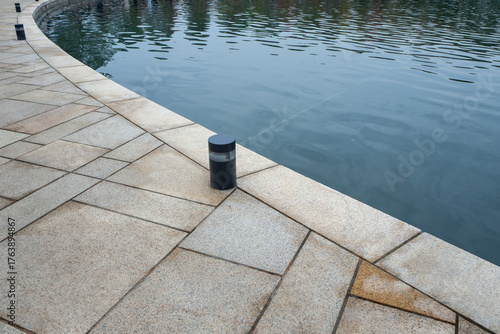 Small cylindrical outdoor light fixture on granite pavement near water surface. Landscape lighting design, waterfront infrastructure, and architectural illumination systems. Real photo.