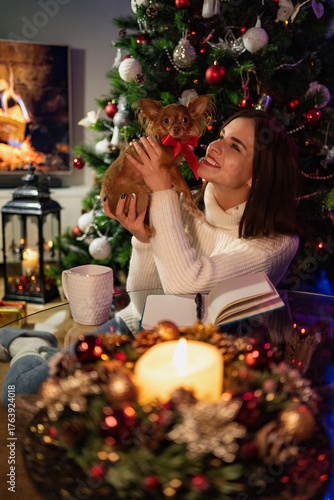 A woman in a white sweater writing in a notebook with a long-haired chihuahua wearing a red ribbon on a glass table, in a Christmas decorated room