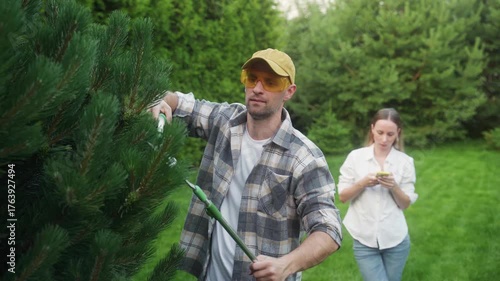 Man in protective glasses and plaid shirt trimming a green pine tree with a long handled pruner in the garden, while a woman in a white shirt and jeans stands behind, engrossed in her mobile phone
