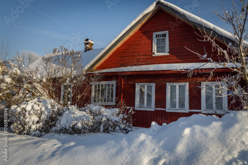 Charming red wooden house blanketed in snow under a crisp blue sky — cozy, picturesque winter scene perfect for holidays, travel, or seasonal lifestyle themes.