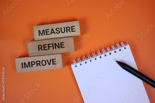 orange background with wooden blocks reading measure, refine, and improve beside a spiral notebook and black pen, symbolizing strategic planning, workflow optimization, and ongoing improvement.