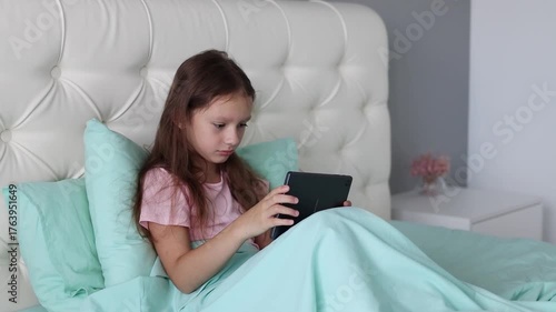 A little girl in pajamas is sitting in the bed after waking up and using a gadget. Problem of excessive phone and tablet use. The concept of a lack of communication in the modern world. Slow motion.
