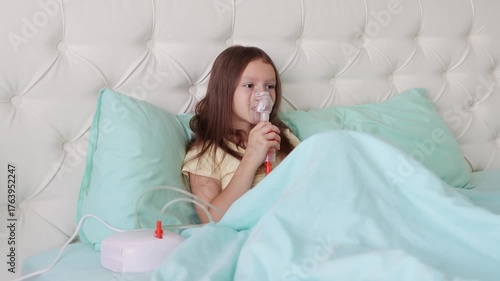 A little girl in pajamas is sitting on a bed and using a nebulizer inhalation mask for respiratory therapy. Child smiling while receiving asthma or cold treatment. 4k footage.