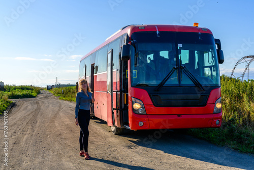 Girl standing beside a red bus on a dirt road during a sunny day in the countryside