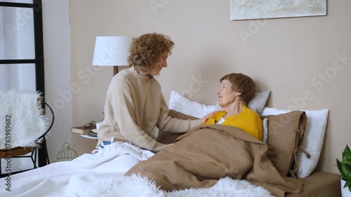 Young adult daughter lovingly cares for her senior mother, tucking her into bed, showing support, affection, and family bonding while having a pleasant conversation in a cozy bedroom