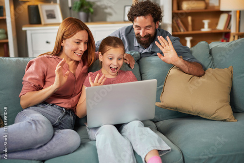 Parents and their daughter share a joyful moment as they interact with loved ones through a laptop, smiling and waving while relaxing on a cozy couch in a stylish living room.