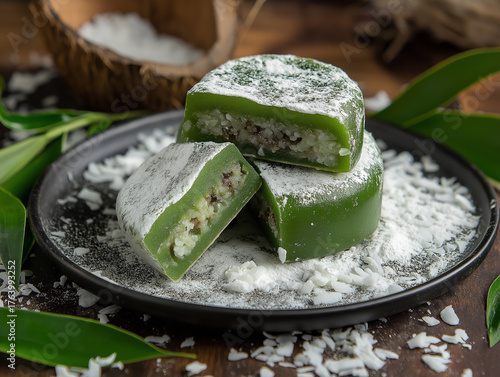 A stack of Pandan Mochi with a deep jade-green tone sits on a dark, matte ceramic plate, surrounded by scattered coconut flakes.