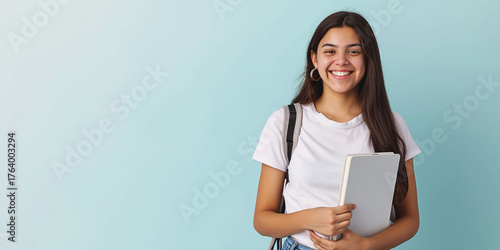 A young Latina teenager, a student, holding a book. She's wearing a white t-shirt.