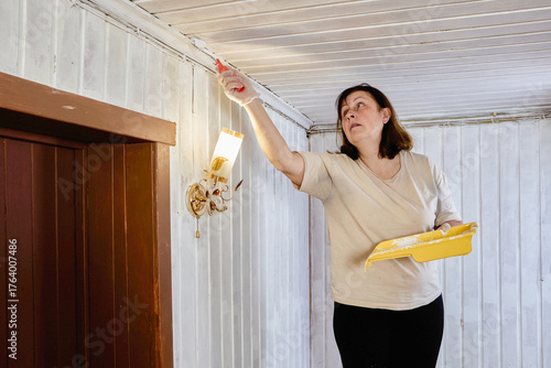 Room undergoes painting to renew surfaces, painter stands on stool applying white paint with roller to ceiling above tongue and groove panels.