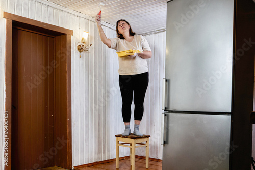 Painter must reach overhead surface to apply white paint, stands on stool and uses small roller to coat ceiling, holds yellow tray above tongue and groove wall.