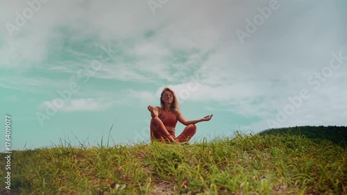 Balanced woman sits in meditation pose at top of hill, eyes closed in serenity. Represents wellness through mindfulness, yoga, and fresh air with natural therapy benefits.