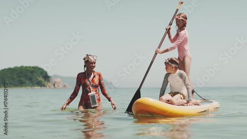 Children playing on paddle board near shore under supervision of trainer, learning and enjoying sunny summer vacation at sea. Concept of carefree childhood and outdoor fun.