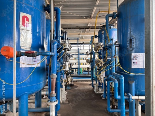 Industrial water treatment plant interior with rows of large blue filtration tanks, complex piping, valves, and control gauges for water purification and chemical processing.