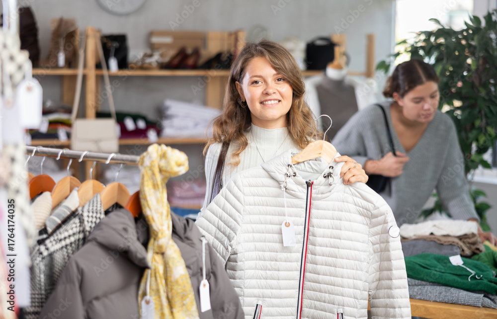 Fototapeta premium Stylish young girl standing in modern clothing shop and looking new down jacket made of high-quality material. Shopping and seasonal sales concept, winter