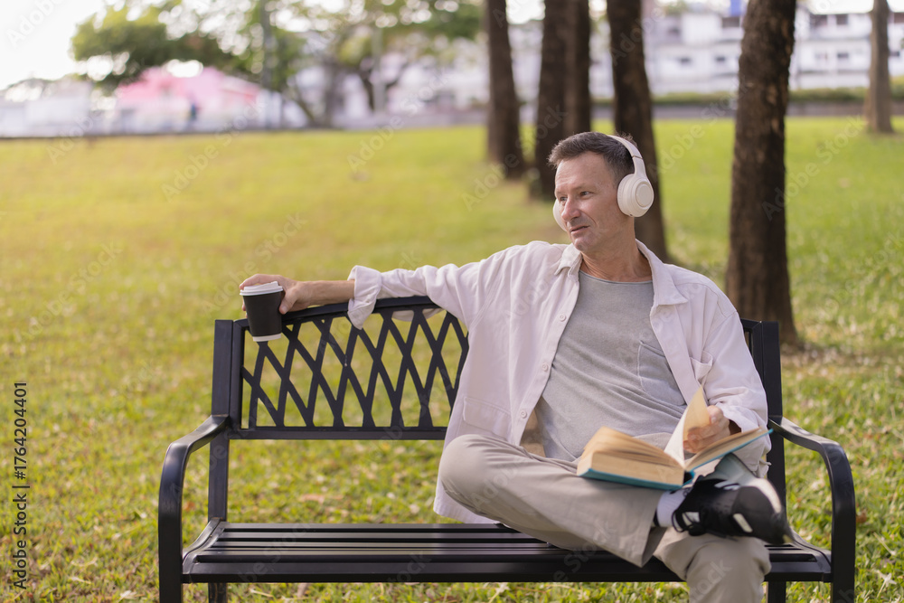 Fototapeta premium Mature man enjoying leisure time on a park bench, listening to music with headphones, holding a coffee cup, and reading a book on a sunny day outdoors