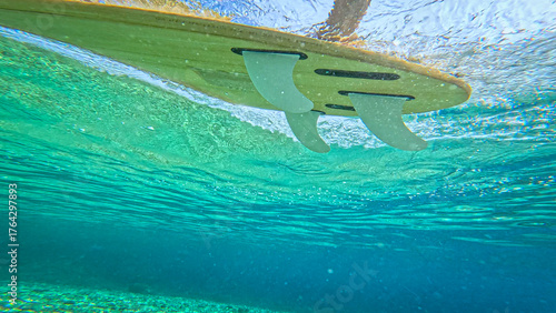 Fotografia UNDERWATER, LOW ANGLE VIEW, CLOSE UP: View of the underside of a wooden surfboard with white fins floating through crystal clear turquoise sea water, leaving behind a ripple of bubbles bellow surface