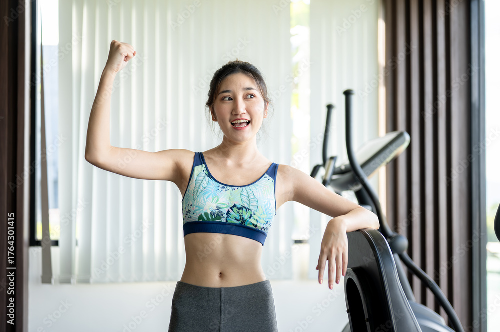 Fototapeta premium Pretty asian woman raising one hand making a fist standing aside exercise equipment in fitness gym.