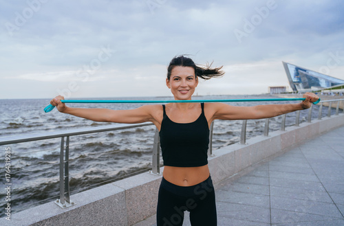 A woman joyfully stretches a resistance band by the ocean, her contagious smile echoing the freedom and vitality of the coastal breeze.
