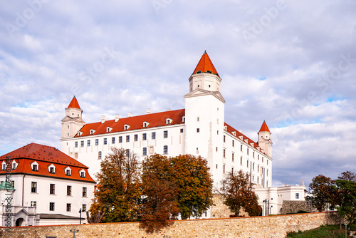 Photography Bratislava Castle in Slovakia with red roofs and white walls, surrounded by autumn trees and flowers under a cloudy sky