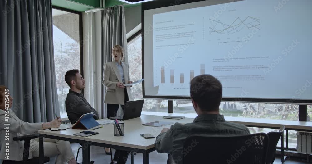 Business team in a meeting room analyzing graphs and charts projected on a screen. A woman leads the presentation, discussing company growth and product performance.