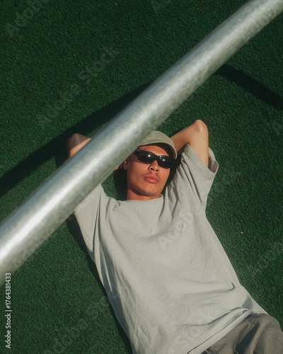 Person relaxes on green surface wearing sunglasses, hat, and casual outfit, enjoying outdoor leisure.
