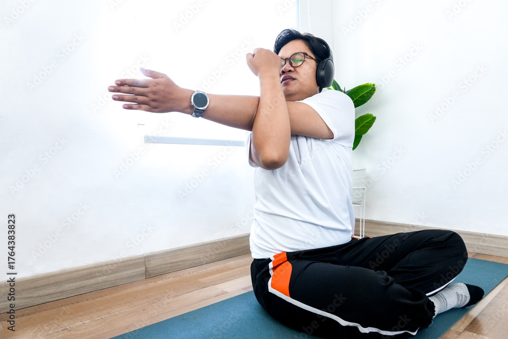 Fototapeta premium Relaxed Young Asian Male Wearing Headphone Stretching Arms During Yoga At Home