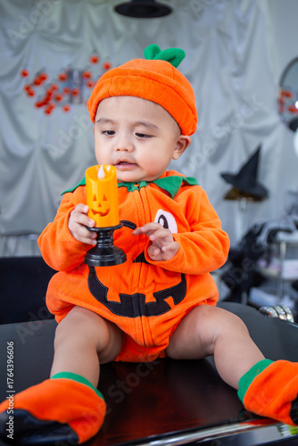 A little boy wearing an orange pumpkin costume and hat sits on a table holding candles to celebrate Halloween.