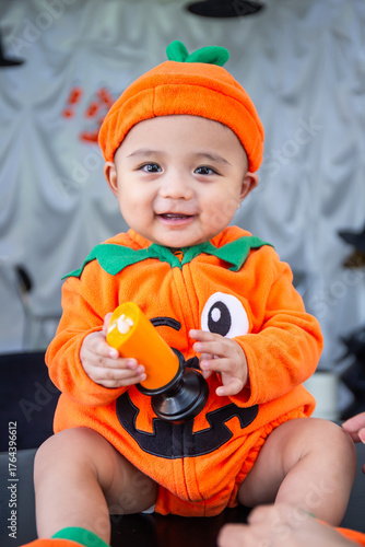 A little boy wearing an orange pumpkin costume and hat sits smiling happily on a table holding candles to celebrate Halloween.