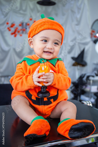 A little boy wearing an orange pumpkin costume and hat sits smiling happily on a table holding candles to celebrate Halloween.
