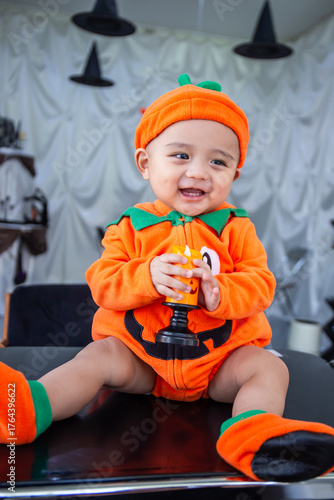 A little boy wearing an orange pumpkin costume and hat sits smiling happily on a table holding candles to celebrate Halloween.