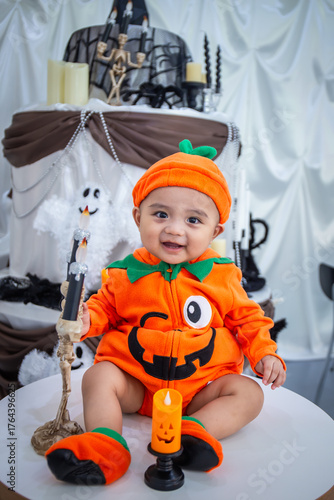 A little boy wearing an orange pumpkin costume and hat sits smiling happily on a table holding candles and skeletons to celebrate Halloween.