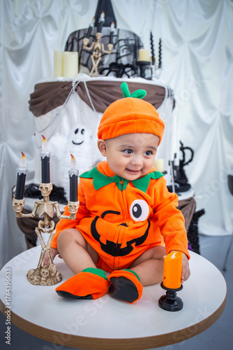 A little boy wearing an orange pumpkin costume and hat sits smiling happily on a table holding candles and skeletons to celebrate Halloween.