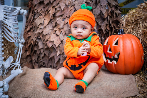 A little boy wearing a pumpkin costume and hat sits on a woven cloth to celebrate Halloween with a background of pumpkins, ghosts and skeletons.
