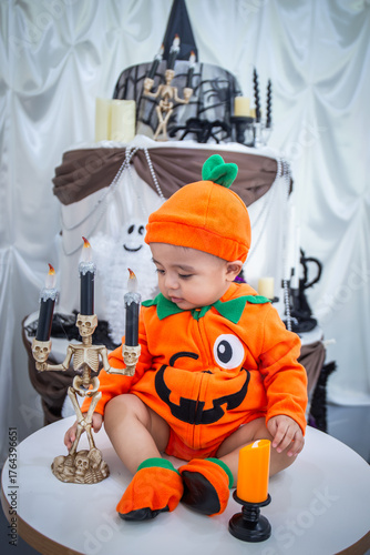 A little boy wearing an orange pumpkin costume and hat sits on a table holding candles and skeletons to celebrate Halloween.