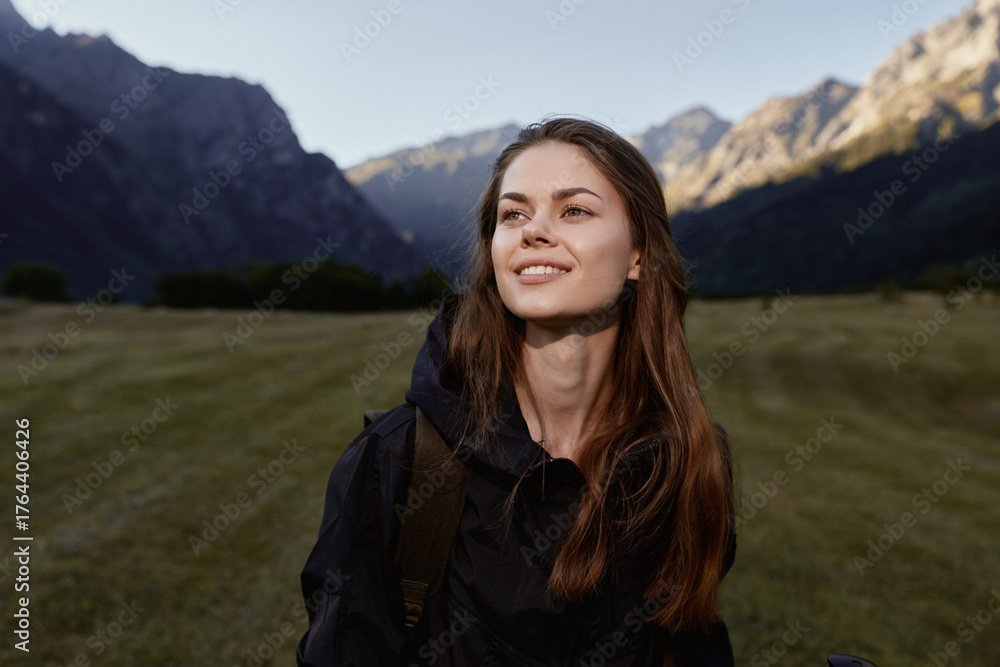 Naklejka premium A cheerful young woman stands outdoors in a scenic mountain landscape. She wears a dark jacket and smiles softly, exuding calm confidence and curiosity as the setting enhances the moment.