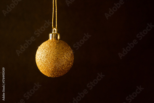 Boule de Noël dorée à paillettes scintillantes suspendue à un fil or à gauche de l'image, arrière plan brun foncé