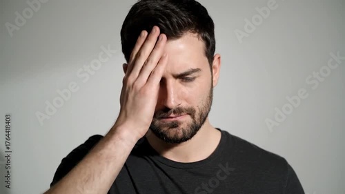 A man with a beard and dark hair holds his hand to his forehead looking down
