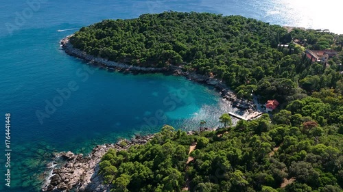 Aerial landscape drone pans left over Lokrum Island’s main port, showing boats docked along rocky shores, dense green trees, and the deep blue Adriatic Sea surrounding the island during autumn.