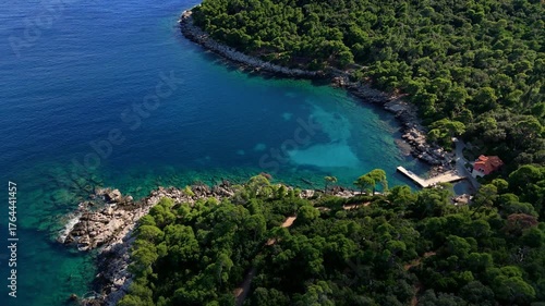 Aerial cinematic landscape drone pans right over Lokrum Island’s port, capturing docked boats, green trees, and the deep blue Adriatic Sea surrounding the rocky coastline of the island.