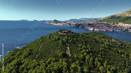 Aerial wide-angle landscape drone flies away from Lokrum Island’s peak, capturing rocky slopes, dense green forest, and the deep blue Adriatic Sea stretching along the island’s coastline in clear day