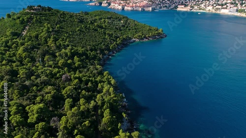 Aerial landscape drone flies right over Lokrum Island, capturing dense green trees surrounded by the vibrant blue Adriatic Sea, with sunlight reflecting off the calm coastal waters and rocky shoreline