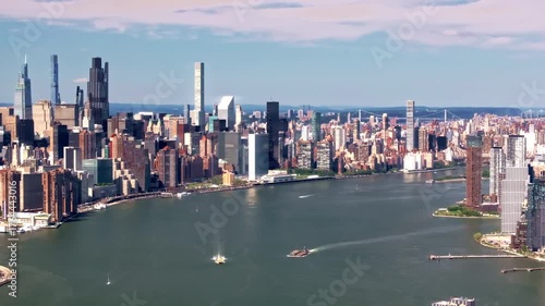 A 70mm cinematic, rotating drone shot over the East River, slowly revealing the Manhattan skyline and its towering skyscrapers, featuring water taxis, and boats traveling up and down the river, of NYC