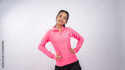 Asian woman wearing a bright pink long-sleeve sports shirt and black shorts standing confidently against a plain white background, smiling with hands on her waist in a cheerful and active posture