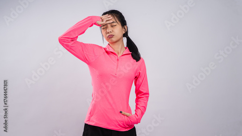 Asian woman in a bright pink long-sleeve sports shirt and black shorts posing against a plain white background, looking tired or stressed with one hand on her forehead and the other in her pocket