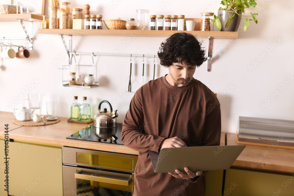 Fototapeta premium Young man engaged in cooking while using a laptop in a cozy kitchen environment