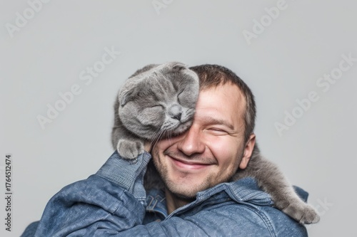 Happy man smiling with gray Scottish Fold cat covering eyes, studio shot, isolated light background, professional photo.