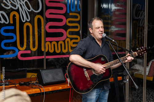 A middle-aged Caucasian man with short brown hair sings into a microphone while playing an acoustic guitar. Colorful background with abstract patterns.