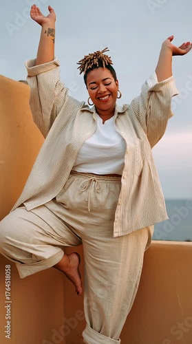 Confident woman smiles while holding a balance pose on rooftop at sunset, expressing freedom and body positivity in peaceful moment.