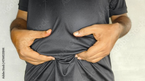 Indian or Asian young adult man's hand holding abdomen or stomach skin and checking excessive belly body fat. Overweight, obesity, weight loss, losing calorie concept. Obese male waist closeup view.