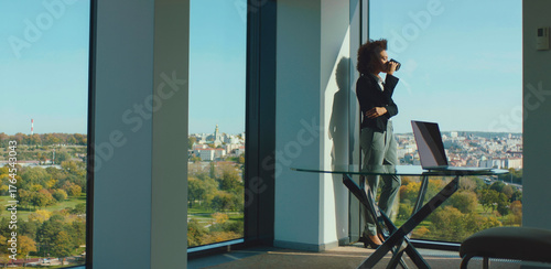 Businesswoman standing by an office window, enjoying a coffee break while observing the expansive city view, a laptop positioned on a modern glass desk nearby, reflecting a moment of contemplation